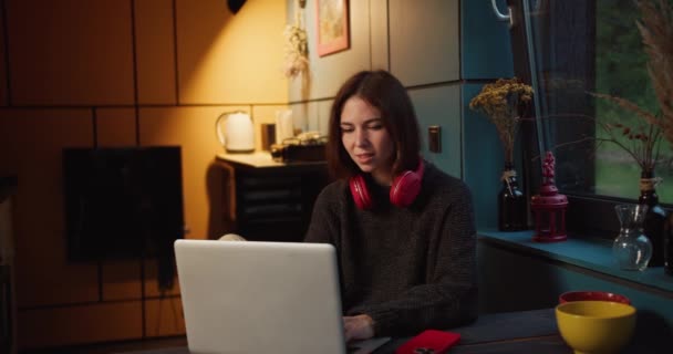 在昏暗的夜晚 一个戴着红色耳机的快乐的黑发女孩一边欣赏着一台白色笔记本电脑的屏幕 一边欣赏着一盏黄色的灯 — 图库视频影像