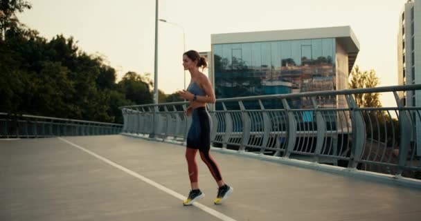 一名身穿夏季运动服的女运动员在城市一座桥的背景下跑步 并在上午跑步前热身 — 图库视频影像