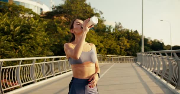 一个穿着夏季运动服的快乐的体育女孩喝着水和一个特殊的瓶子 然后把它倒在她的脸上 累了的女孩慢跑后洗脸 — 图库视频影像