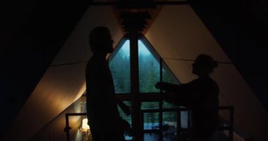 Bir adamla bir kızın siluetleri, karanlık bir odada yeşil bir dağ ormanına bakan bir pencerenin arka planına sarılıyorlar. romantik buluşma.