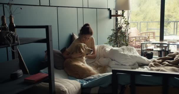 一个穿着浅色T恤的黑发女孩躺在床上 在家里和她那只浅色的大狗在一道碧绿的墙后面玩耍 — 图库视频影像