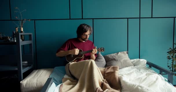 暗い赤いシャツのブルネット男は 毛布に包まれたソファの上に横たわり ターコイズの壁に対してギターを演奏します — ストック動画