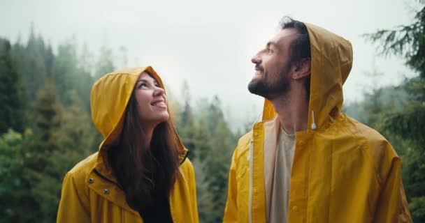 一对快乐的夫妇 一个穿着黄色夹克的男人和一个女孩站在雾蒙蒙的山林中 微笑着等待即将到来的雨 男孩和女孩看着天空 拥抱着 — 图库视频影像