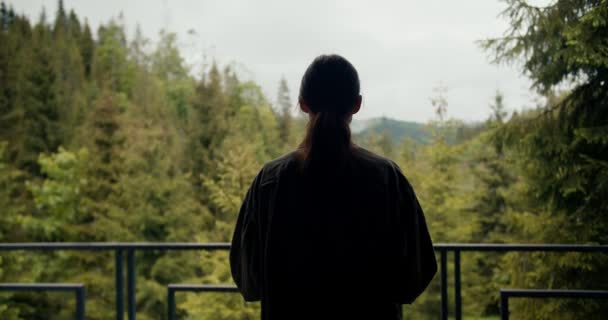 一个穿着绿夹克 头戴绿茶的快乐的黑发女孩走到一座俯瞰高山上一片绿针叶林的乡间房子的阳台上 — 图库视频影像