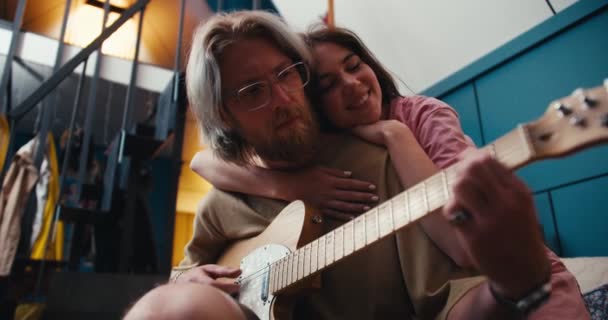 一个戴着长发眼镜 留着胡子的金发姑娘弹电吉他 一个黑发姑娘抱着他的肩膀 听着他在舒适的房间里弹奏 — 图库视频影像