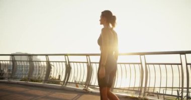 Spor üniformalı bir kız gün doğumunun arka planında köprü boyunca yürüyor..