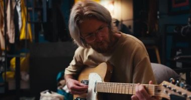 Uzun saçlı, sakallı, sarışın bir adam rahat bir odada elektro gitar çalıyor..