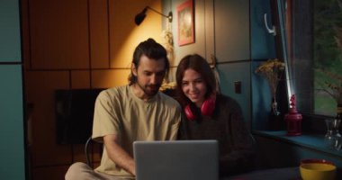 Esmer bir adam ve kırmızı kulaklıklı esmer bir kız akşam üstü dizüstü bilgisayarda oturup arka planda sarı bir lamba olan bir video izlerler..