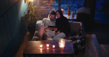Romantik bir akşam: esmer bir adam ve bir kız kanepede oturuyor ve akşamları tabletin ekranına bakıyorlar. Masanın üzerinde mumlar var..