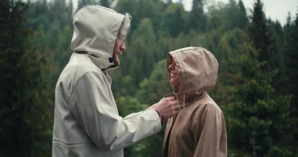 男人把女孩们的外套系好 在多山的针叶林里在雨中亲吻她 绿林背景下的快乐夫妻 — 图库视频影像