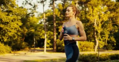Spor üniformalı bir kız sabah parkında koşuyor ve siyah bir şişeden üzerine su döküyor..