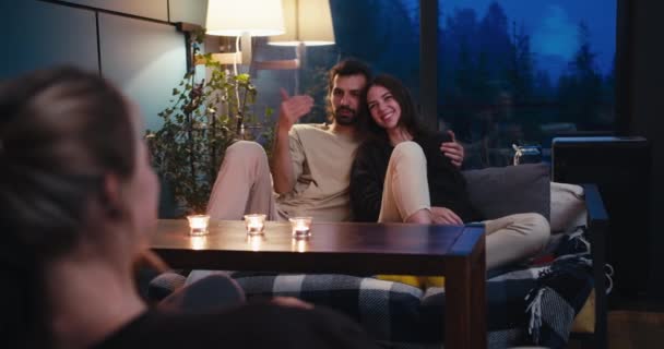 一个男人和一个女孩坐在沙发上 和一个金发姑娘在桌上聊天 桌上有蜡烛 晚上朋友们舒服的交谈 — 图库视频影像