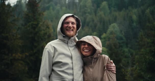 ハッピーカップル ヒゲとジャケットのブロンドの少女が立って 山の緑の森の背景に喜んでメガネでブロンドの男 — ストック動画