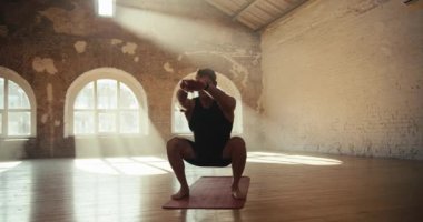 Sunny Hall 'da siyah spor üniformalı genç bir adam özel bir halıda spor yapıyor. Bacak kaslarının gelişmesi için çömelmeler ve diğer egzersizler.