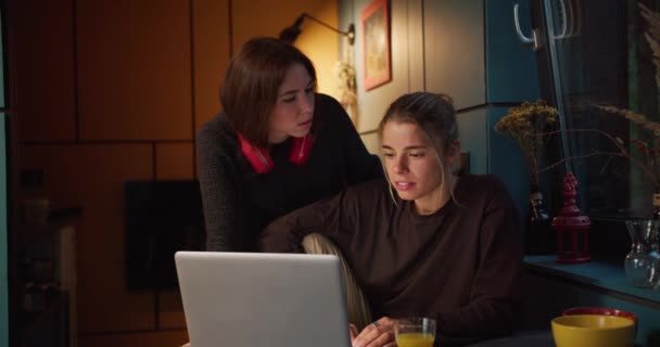 傍晚时分 一个黑发女孩和一个金发女孩在一间黑暗的房间里玩乐地望着一台白色笔记本电脑的屏幕 屏幕上亮着一盏神圣的黄灯 — 图库视频影像