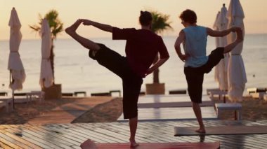İki adam sahilde tek ayak üzerinde meditasyon yapıyor. Sabah plajda Zen tarzı yoga dersi..