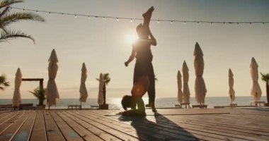 Adam arkadaşının başının üstünde özel bir halıya çıkıp bacaklarını desteklemesine yardım ediyor. Yoga dersleri ve sabah egzersizleri birlikte..