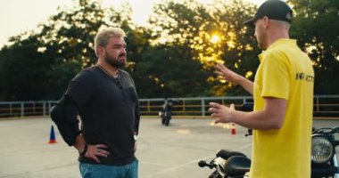 Gri kazaklı ve gür sakallı bir adam sarı tişörtlü bir sürücü eğitmenini dikkatle dinler. Motosiklet sürmeden önce eğitim.