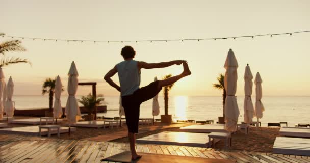 一个年轻人抬起腿 单腿站在阳光灿烂的海滩上的后视镜 日出日落时的瑜伽和伸展课 — 图库视频影像