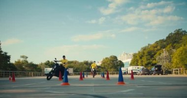 Sarı tişörtlü iki sürücü eğitmeni motosiklet manevra kabiliyetlerini geliştiriyor. İki adam motosikletle engelleri aşıyor..