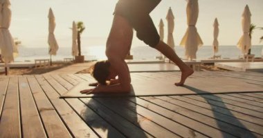 Adam güneşli bir sahilde özel bir paspasın üzerinde başının üstünde duruyor. Yoga dersleri.