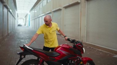 Yan görüş. Sarı tişörtlü kel bir adam kırmızı bir motosikleti garaja sürüyor. Motosiklet okulunda günün sonunda..