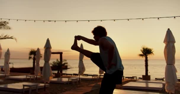 一个年轻人抬起他的腿 在早上进行一场阳光灿烂的促销活动 早上在桑尼海滩做运动和瑜伽 健康的生活方式 — 图库视频影像