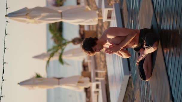 黒いショートパンツの男がマットの上に座って 朝のビーチでヨガをする 瞑想クラスは心を落ち着かせる 身体と心の調和 — ストック動画