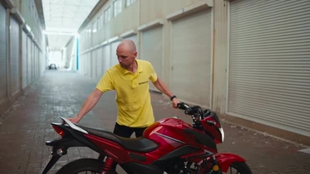 侧视图 一个穿着黄色T恤的秃头司机驾驶一辆红色摩托车进入车库 在摩托车学校的工作日结束 — 图库视频影像