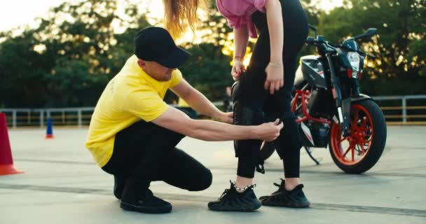 一个穿着黄色T恤的驾驶学校的教师帮助一个女孩在骑摩托车时戴上防护板 骑摩托车时的脚垫和其他防护措施 — 图库视频影像