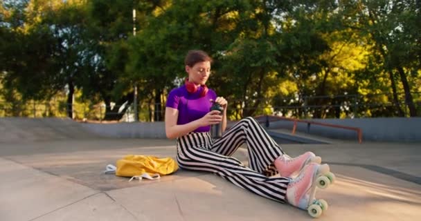 一个有着紫色上衣和粉色轮滑鞋条纹裤子的短发女孩坐在混凝土地板上的一个滑板公园里 喝着一瓶特制的水 — 图库视频影像