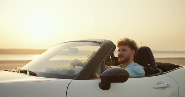 青いシャツのカーリーヘアのひげ付き男は 川の近くのフィールドを横切って白いコンバーチブルカーで彼のブロンドのガールフレンドを運転します — ストック動画