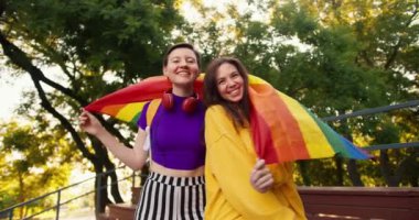 Mor bataklıkta kısa saçlı bir kız ve sarı kazaklı esmer bir kız kendilerini LGBT topluluğunun bayrağının altına sarmışlar..