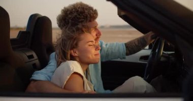 Kıvırcık saçlı sakallı bir adam sarışın kız arkadaşına üstü açık bir arabada sarılıyor ve rüzgarlı bir havada arka planda birlikte dört gözle bekliyorlar..