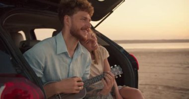 Mavi gömlekli, kıvırcık saçlı sakallı bir adam ukulele çalıp şarkı söylerken sarışın kız arkadaşı başını omzuna yaslıyor. Gülümsüyorlar ve arabanın bagajında oturuyorlar.