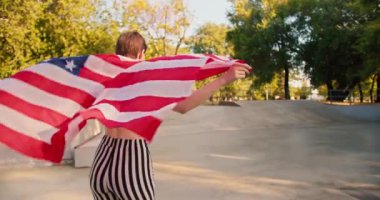 Kırmızı kulaklıklı, mor tişörtlü, kısa saçlı mutlu bir kız kaykay parkında paten kayıyor ve elinde ABD bayrağını tutuyor..