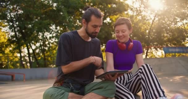 一个穿着灰色T恤的黑发男人和一个穿着紫色上衣剪短头发的女孩在公园里看平板屏幕上的东西 — 图库视频影像