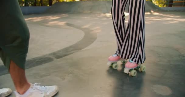 一个穿着灰色T恤的黑发男子在混凝土地板上的溜冰场帮助一个身穿紫色上衣和条纹裤子的女孩骑4轮粉色轮滑冰鞋 — 图库视频影像