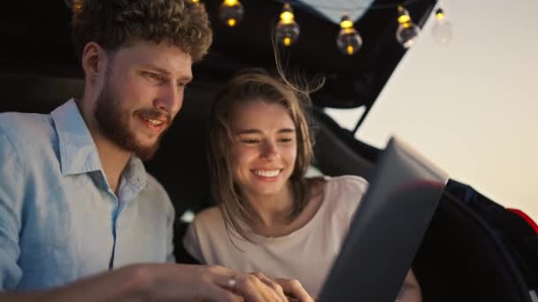 一个留着胡子 留着一头卷发的蓝色衬衫的男人 在用灯泡装饰的一辆黑色轿车的行李箱里 向他的女朋友展示了笔记本电脑的屏幕 女朋友正靠着他笑着笑着笑着 — 图库视频影像