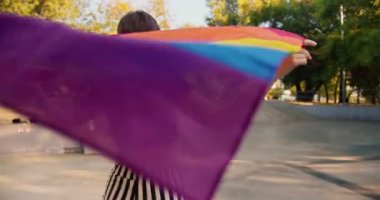 Paten parkında paten kayan ve rüzgarda dalgalanan bir LGBT bayrağı tutan mor bluzlu ve çizgili pantolonlu mutlu bir kızın portresi..