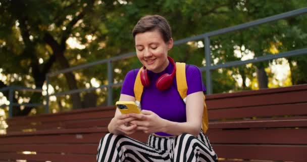 一个穿着紫色上衣 条纹裤子和黄色背包的短发女孩坐在棕色的架子上 用黄色的电话聊天 — 图库视频影像