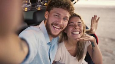 Mavi tişörtlü, kıvırcık saçlı sakallı bir adam sarışın kız arkadaşıyla kameraya gülümserken siyah bir arabanın bagajında ampullerle süslenmiş selfie çekiyor..