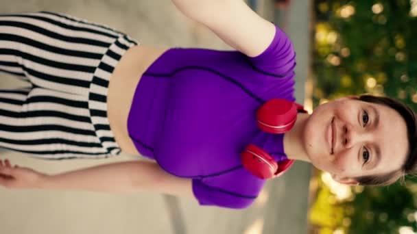 垂直视频 快乐的女孩 头上戴紫色红色耳机 穿着条纹裤子 骑着溜冰鞋在公园里短短的理发 还带着一个自拍标志V — 图库视频影像