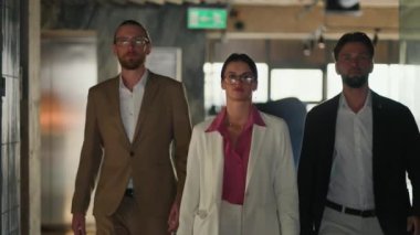 Ofis çalışanlarından oluşan kendinden emin bir üçlü, kahverengi ceketli sarışın bir adam, pembe gömlekli esmer bir kız ve mavi ceketli bir adam modern ofis boyunca güvenle yürüyorlar..