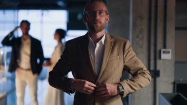 Sarışın ve sakallı bir adamın portresi açık kahverengi takımını ayarlıyor, kollarını göğsüne koyuyor, gülümsüyor ve modern bir ofise karşı kameraya bakıyor..