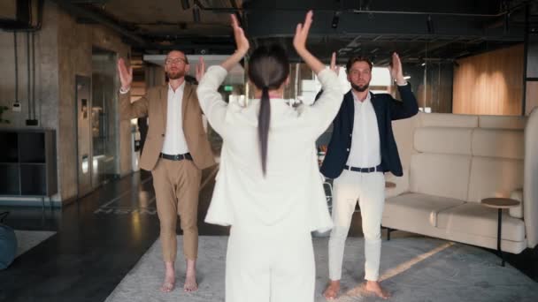 在一个现代化的办公室里上瑜伽课 一名身穿白色西服的女孩和她的同事们 穿着棕色和蓝色夹克的小伙子们 在一个现代办公室的垫子上上瑜伽课 站在一个特殊的姿势上 — 图库视频影像