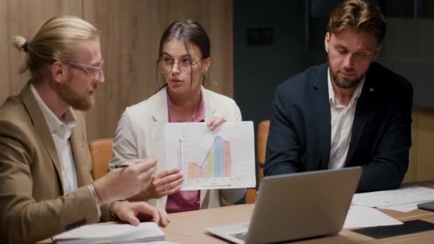 ピンクのシャツと丸い眼鏡の白いスーツのブルネットの少女は 現代のビジネスススーツの2人の男の会社のビデオ会議で彼女の対話者に紙のシート上のグラフを示しています — ストック動画
