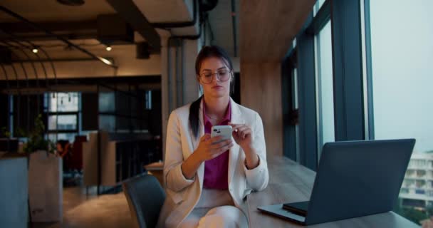 一个身穿白色夹克和粉色衬衫 戴着圆形眼镜的黑发女孩的画像 她在一间现代化办公室的全景窗旁边的一部白色电话里工作 — 图库视频影像