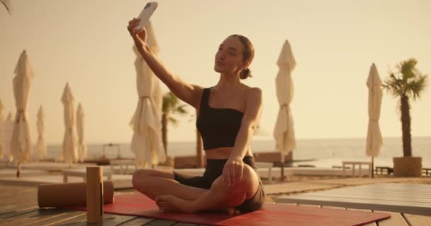 一个黑头发的黑发女孩在沙滩上拿起一个带着雨伞和日光浴床的红色垫子上的白色手机拍了个自拍 — 图库视频影像