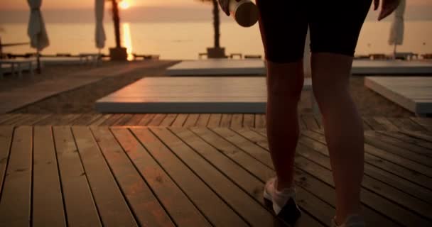 一名身穿黑色运动服的女孩在黎明时分身穿黑色运动服 带着木板和瓶装水沿着海滩散步 准备早上的体育活动 在日出时的冥想 — 图库视频影像
